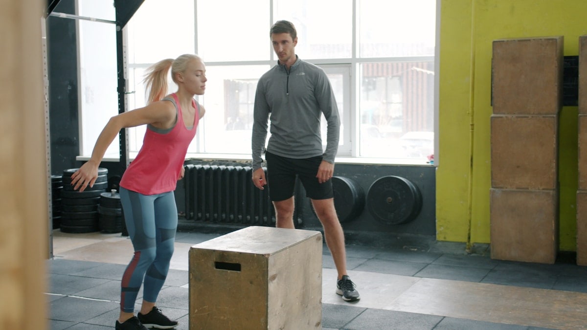 Girl in sports clothing is jumping on wooden box training while sports coach is watching standing nearby. People, well-being and aerobic exercises concept.